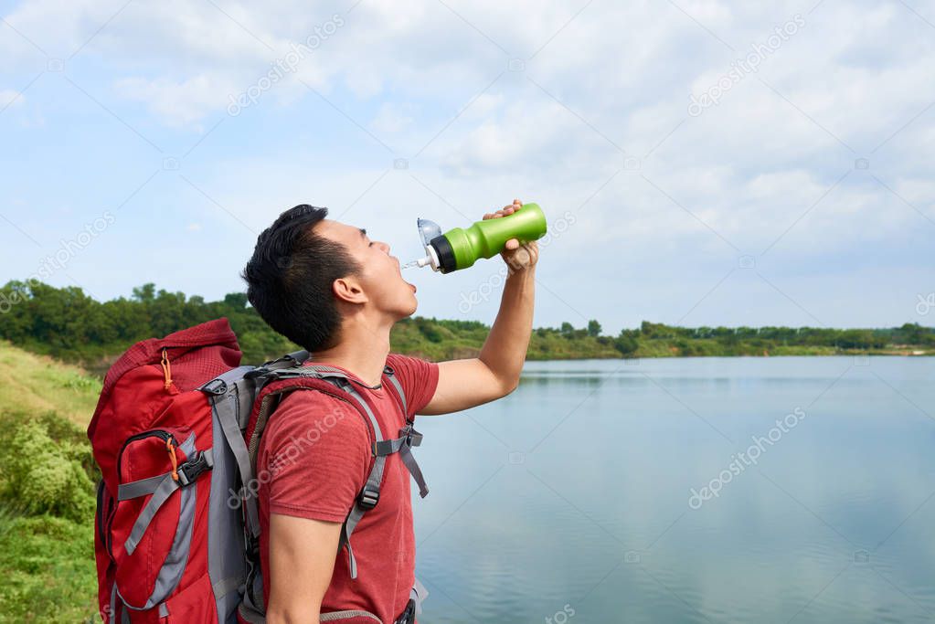 Young thirsty traveler drinking water after hiking with big backpack