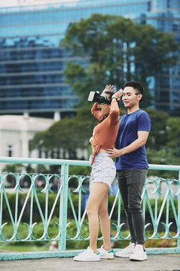 Heyecanlı genç kadın şehir açık havada ayakta iken onu destekleyen genç adam ile birlikte sanal gerçeklik oyunu oynuyor