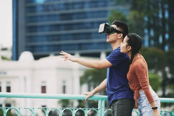 Young man looking at virtual reality goggles and gesturing with his girlfriend standing behind him in the city