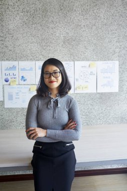 Arka planda mali grafikler ile ofiste duran gözlük Asyalı genç iş kadını Portresi