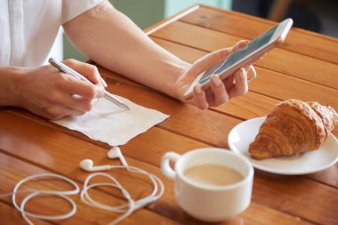 Hands of woman checking her phone and writing details on napkin at cafe table clipart