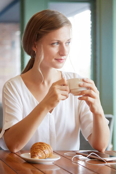 Pensive attractive young woman enjoying cup of tea and croissant for breakfast