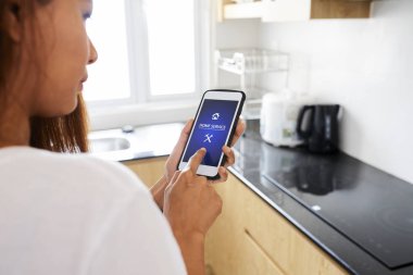 Akıllı telefonu olan genç bir kadın ev mutfağında dururken ev servisi uygulamasını kullanıyor.