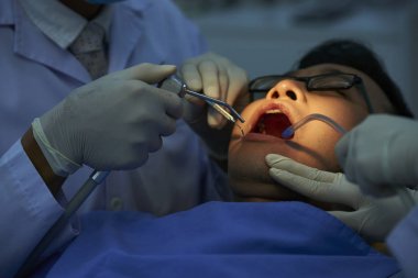 İki diş hekimi dişlerini kontrol edip klinikte delerken ağzı açık yatan erkek hasta