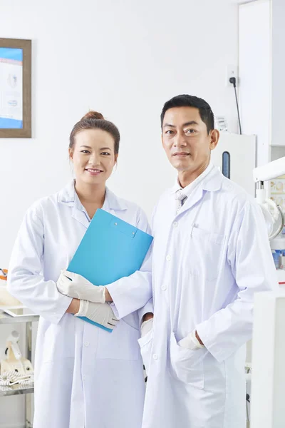 歯科医院のクリップボードを持つ女性看護師と一緒に立っている白いコートの成熟した男性歯科医の肖像画 — ストック写真
