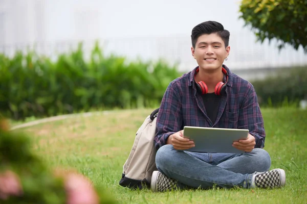 微笑的亚洲大学生与穿孔的耳朵坐在绿色草坪上与平板电脑 — 图库照片