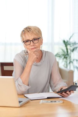 Pensive kıdemli kadın dizüstü bilgisayar ve not defteri ile masada oturan ve onun mali hesaplama