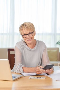 Dizüstü bilgisayar ve not defteri önünde masada oturan ve hesap makinesi ile onun mali sayma olgun kadın Portresi