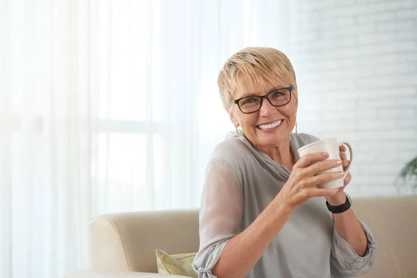 コーヒーを飲みながらソファに座り 自宅のカメラで微笑む眼鏡をかけた幸せな成熟した女性の肖像画 — ストック写真