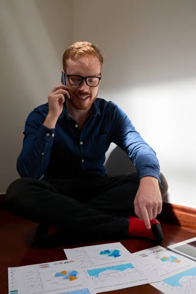 Smiling young financial adviser sitting on the floor analyzing financial graphs and charts and consulting somebody on mobile phone