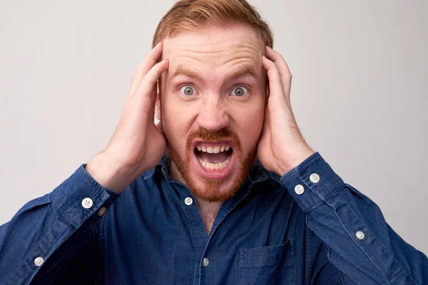 Portrait of red haired young man in panic holding his head and looking at camera