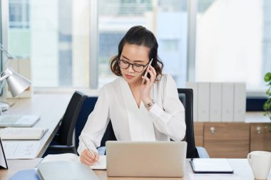 Gözlüklü güzel genç Vietnamlı iş kadını telefonda konuşuyor ve not alıyor 