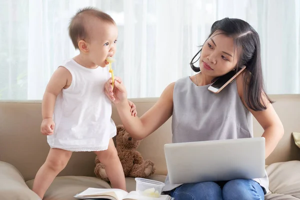 電話でクライアントに電話をかけるときに彼女の小さな赤ちゃんを養うフリーランスの女性 — ストック写真