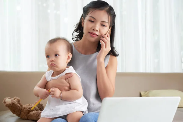 彼女のラップトップ上の赤ちゃんと一緒に座って 電話で呼び出すかなりベトナムの女性 — ストック写真