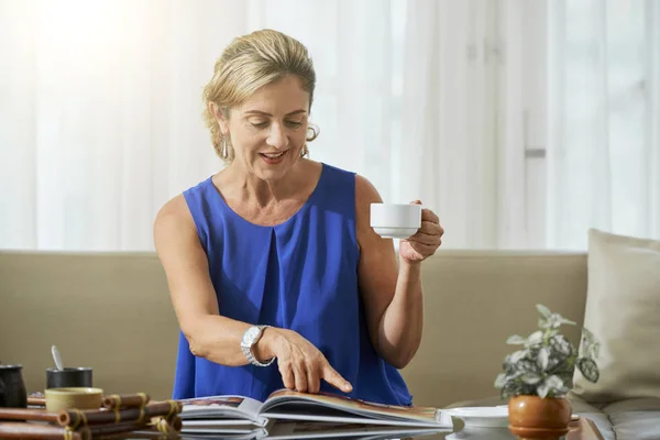 自宅のフォトアルバムの写真を指し示すお茶のカップでソファに座っている成熟した女性 — ストック写真
