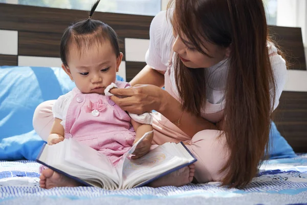Genç Asyalı kadın ilginç bir kitap okuyan küçük kızının yüzünü siliyor.