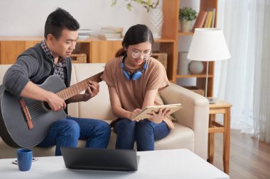 Yakışıklı Asyalı adam akustik gitar çalmak ve yakındaki ışık kanepede oturan gözlük muhteşem gülümseyen genç kadın tarafından düzenlenen tablet bakarak