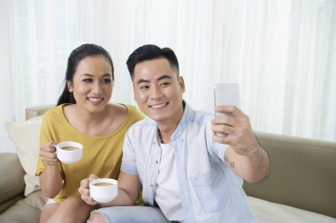 Neşeli genç Vietnamlı çift kahve içiyor ve evde akıllı telefon selfie alıyor