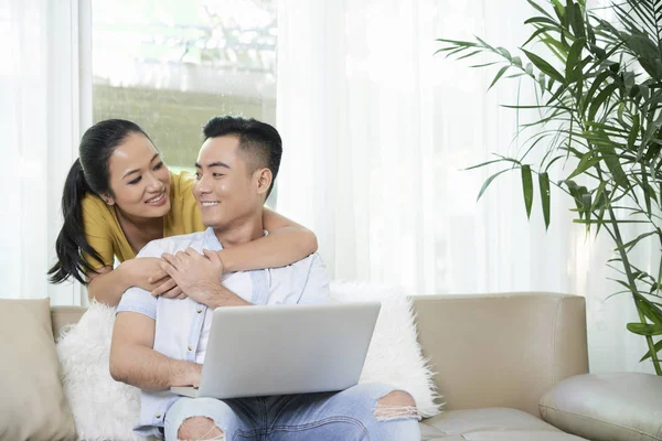年轻漂亮的亚洲女人在丈夫在家用笔记本电脑的时候 从背后拥抱他 — 图库照片