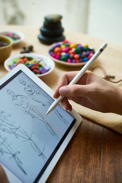 Close-up of woman sitting at wooden table and drawing sketches of clothes for female on digital tablet