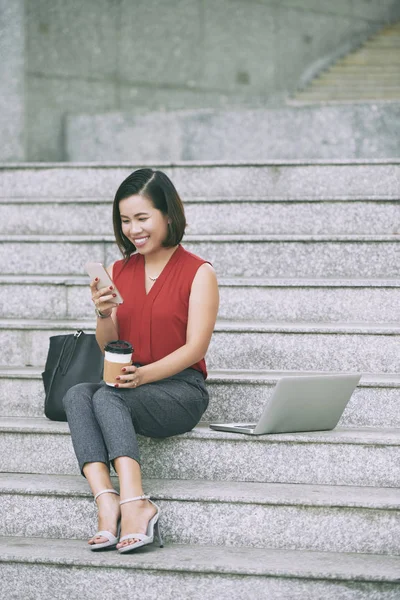 石の階段に座るアジアの幸せなビジネスウーマンは 笑顔で携帯電話でメッセージを読み コーヒーマグを持っています — ストック写真