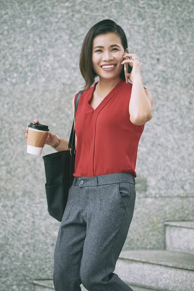 可処分のコーヒーを持ち 屋外の階段を歩きながら携帯電話で話すアジアのビジネスウーマン — ストック写真