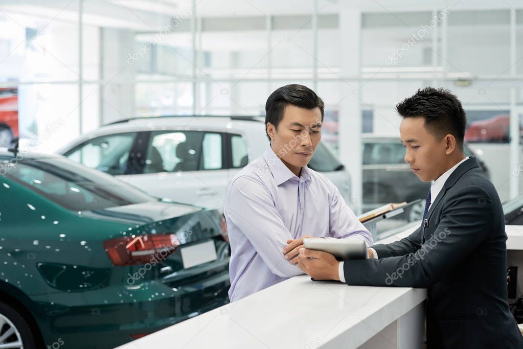 Young professional sales manager of car dealership showing contract details on tablet computer to customer