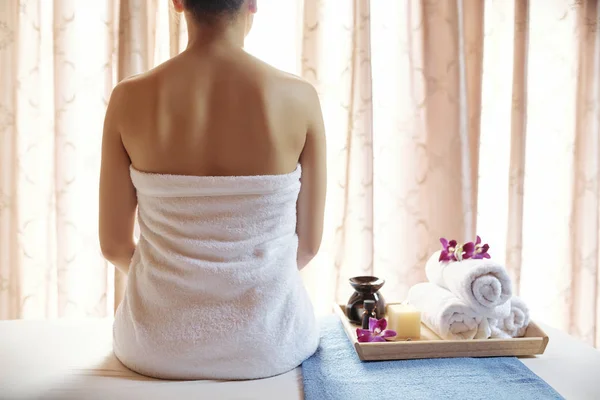 Rear view of young woman wrapped in towel sitting on bed in luxurious spa salon