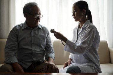 Beyaz önlüklü Asyalı kadın doktor, evde kanepede otururken üst düzey hastasının kalbini dinleyecek.