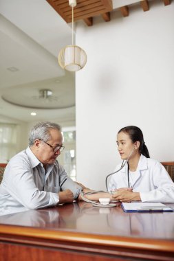 Asyalı kadın doktor evde kan basıncını kontrol ederken gözlük kıdemli adam masada otururken