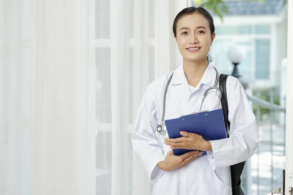 青いフォルダーを持ち 患者の家を訪問しながらカメラを見ている白いコートに立つ若いアジアの看護師の肖像画 — ストック写真