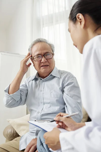 メガネをかけた先輩男性が若い看護師に頭痛を訴え 家を訪れる際に処方箋を書いている — ストック写真