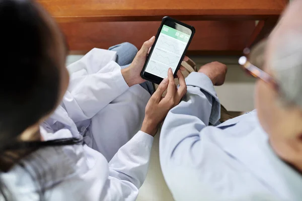 Close-up of female doctor holding mobile phone and showing how to use mobile application to controlling the health to senior man sitting near by her