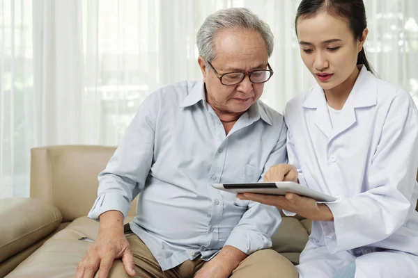 彼らはソファに一緒に座っている間 シニア患者にデジタルタブレット上のオンライン治療を示すアジアの女性医師 — ストック写真