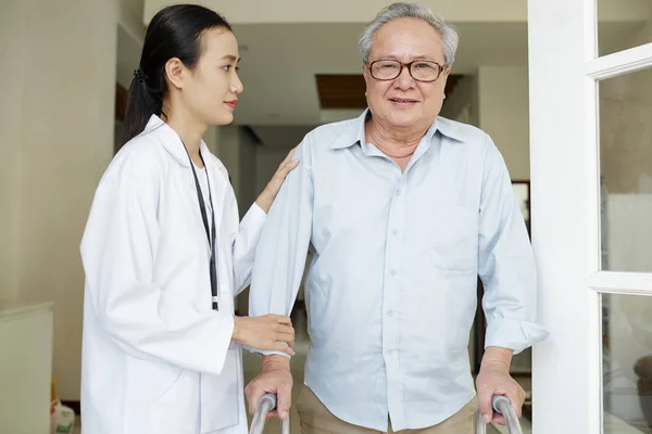 Old man in eyeglasses standing with walker and looking at camera while nurse in white coat helping him to move