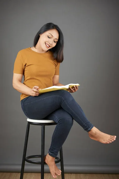 穿着牛仔裤和T恤坐在凳子上看小说的年轻而积极的亚洲美女 — 图库照片