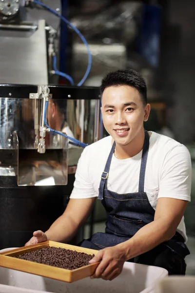 亚洲年轻大师在生产咖啡工厂工作期间 坐在筛子上 拿着新鲜的咖啡豆 对着镜头微笑 — 图库照片