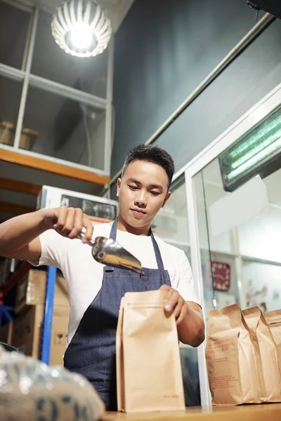 アジアの若者がエプロンに立ち 生産後に販売のための焙煎コーヒー豆で紙袋を充填 — ストック写真