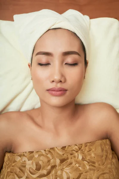 Portrait of Asian young woman with towel on her head lying with eyes closed and relaxing in spa salon