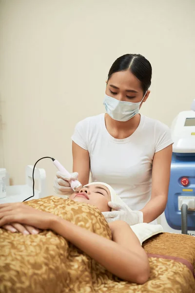 Asian specialist in protective mask standing and doing ultrasound cleaning of woman face in cosmetologist cabinet