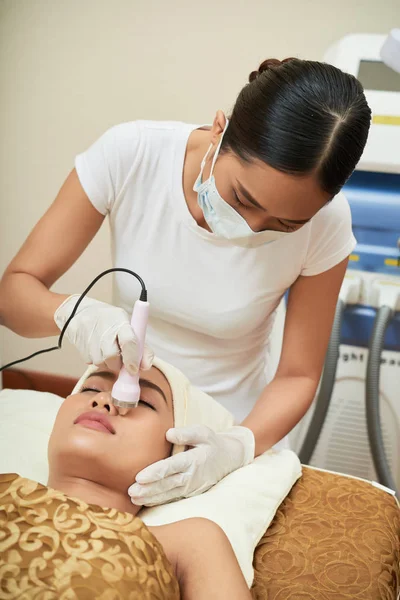 Asian young specialist doing cleaning of face with ultrasound while woman relaxing with eyes closed in spa salon