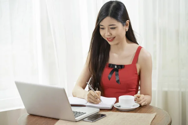 面带微笑的亚洲女商人坐在桌旁 在记事本上做咖啡做笔记 看着电脑笔记本电脑的显示器 — 图库照片