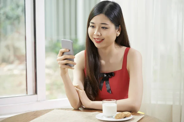 美丽的亚洲女人坐在桌旁喝着牛奶 喝着饼干 用手机打信息 — 图库照片