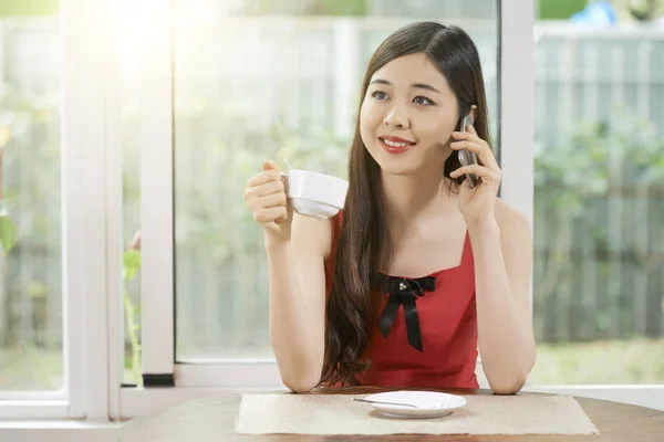 Menina Asiática Sentada Mesa Conversando Telefone Celular Sorrindo Enquanto Bebe — Fotografia de Stock