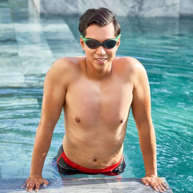 Yüzme gözlüğü takan ve açık yüzme havuzunda yüzerken kameraya gülümseyen genç gömleksiz adamın portresi