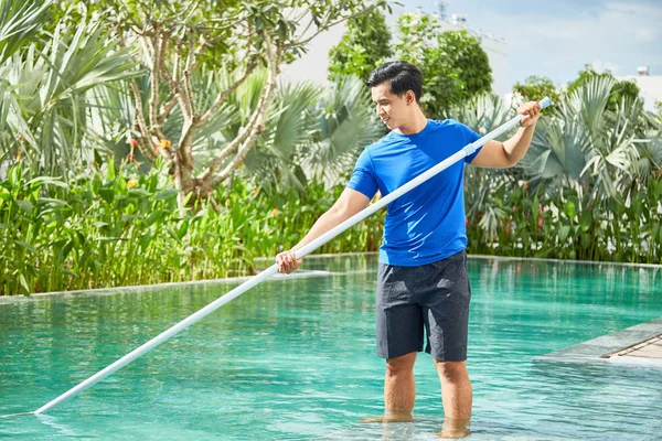 アジアの若い男性クリーナーは 水の中に立って 屋外の特別な機器でスイミングプールを掃除 — ストック写真