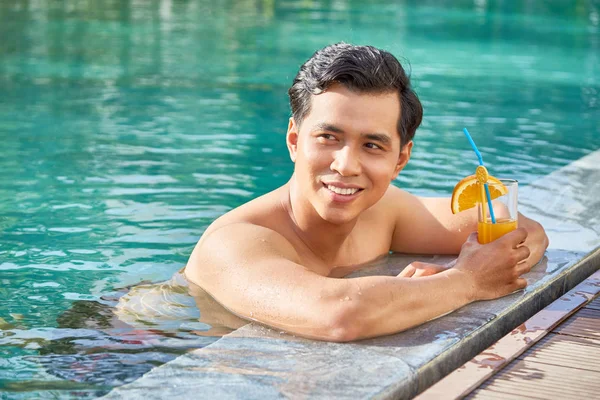 Asian smiling man relaxing in the water at poolside with glass of orange juice at resort during summer vacation