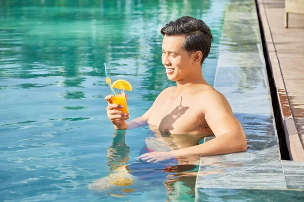 年轻快乐的男子坐在池塘边的水里 在度假胜地欣赏着一杯橙汁 — 图库照片