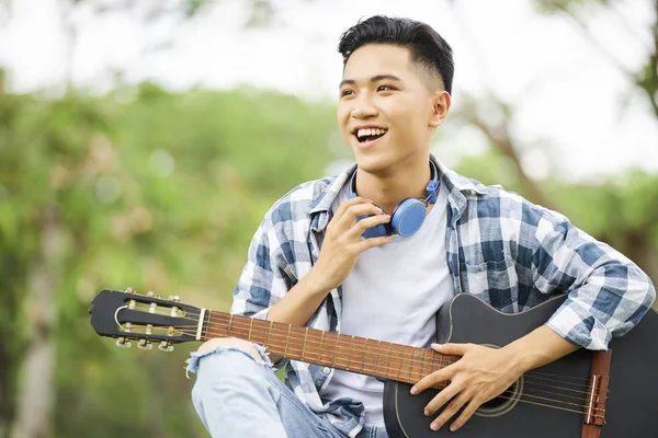 カジュアルな服を着て 公園でギターで座っている彼の首にヘッドフォンでアジアの笑顔ティーンエイジャー — ストック写真