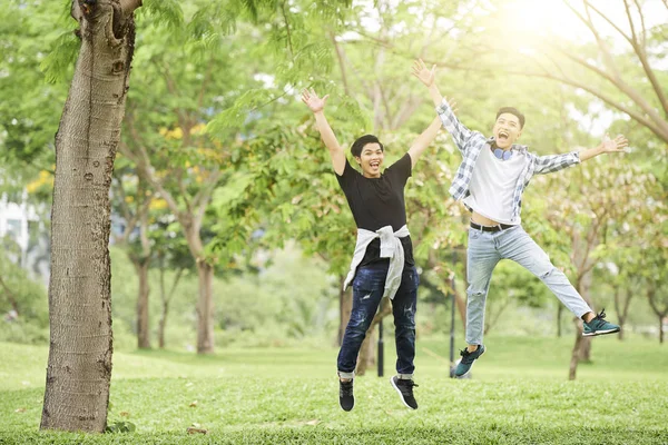 カジュアルな服を着た2人の興奮したティーンエイジャーの肖像画は 公園で一緒にジャンプし カメラで微笑んでいます — ストック写真